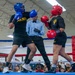 Two U.S. Army soldiers are competing in a boxing match inside what appears to be a military facility or gymnasium. A referee dressed in a light blue shirt and bow tie oversees the bout as one boxer in blue gear throws a punch at the opponent in red gear. A large crowd of military personnel and spectators watches from the background.