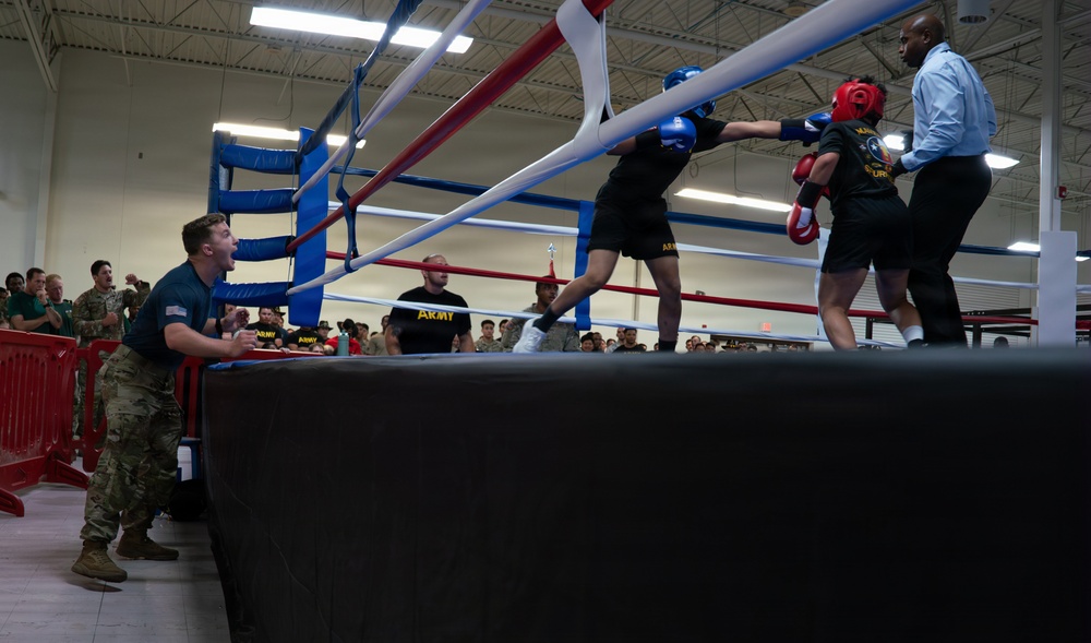 A military boxing match is taking place inside an indoor gymnasium or sports facility. Two boxers wearing protective headgear and gloves are engaged in a bout, overseen by a referee in a blue shirt, while a soldier in camouflage uniform shouts instructions or encouragement from ringside. A crowd of military personnel in Army uniforms watches from the background.