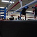 A military boxing match is taking place inside an indoor gymnasium or sports facility. Two boxers wearing protective headgear and gloves are engaged in a bout, overseen by a referee in a blue shirt, while a soldier in camouflage uniform shouts instructions or encouragement from ringside. A crowd of military personnel in Army uniforms watches from the background.