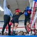 A military boxing match is taking place inside an indoor arena, with two soldiers wearing Army-branded shorts exchanging punches in close range. A referee in a light blue dress shirt and black pants has his arm raised, appearing to intervene or signal a stoppage. A large crowd of military personnel in uniform watches from ringside.