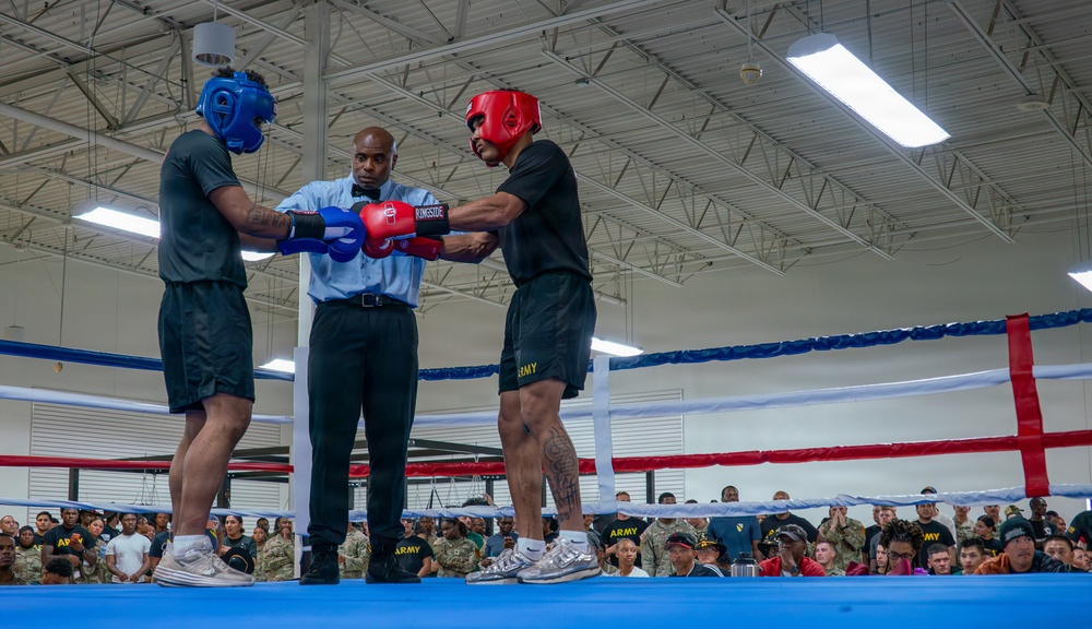 Two boxers wearing protective headgear and gloves are being brought together by a referee in a boxing ring, likely at the start of a bout or during a pre-fight instruction. Both fighters are wearing black shorts with 'ARMY' printed on them, suggesting this is a military boxing event. A large crowd of spectators, many in military uniforms, watches from ringside in what appears to be an indoor military facility.