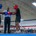 Two boxers wearing protective headgear and gloves are being brought together by a referee in a boxing ring, likely at the start of a bout or during a pre-fight instruction. Both fighters are wearing black shorts with 'ARMY' printed on them, suggesting this is a military boxing event. A large crowd of spectators, many in military uniforms, watches from ringside in what appears to be an indoor military facility.