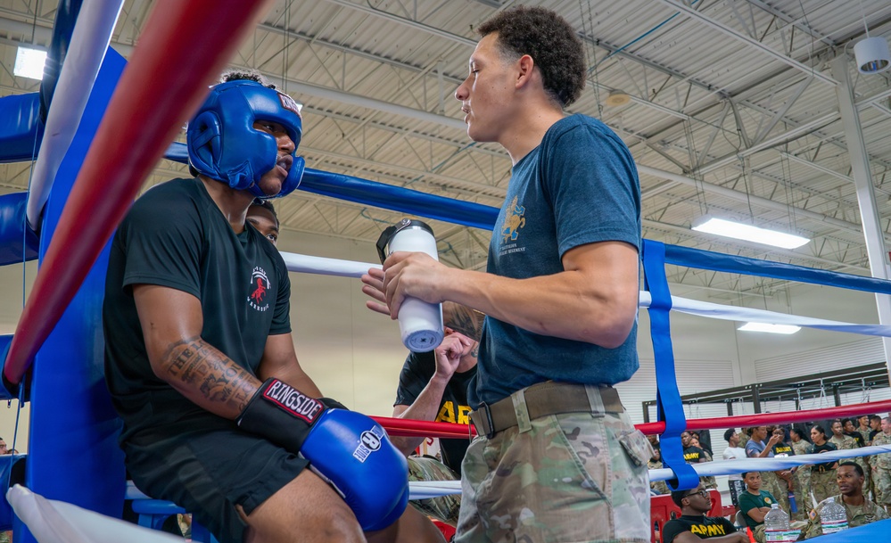 A boxing match corner scene showing a fighter seated on a stool in the ring corner between rounds, receiving attention from a corner person/trainer. The fighter is wearing blue Ringside boxing gloves and a blue headguard, while the trainer in a blue t-shirt and camouflage pants holds what appears to be a water bottle or medical supply. The event appears to be a military boxing competition, evidenced by the Army branding, uniformed spectators, and indoor gymnasium setting.