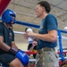 A boxing match corner scene showing a fighter seated on a stool in the ring corner between rounds, receiving attention from a corner person/trainer. The fighter is wearing blue Ringside boxing gloves and a blue headguard, while the trainer in a blue t-shirt and camouflage pants holds what appears to be a water bottle or medical supply. The event appears to be a military boxing competition, evidenced by the Army branding, uniformed spectators, and indoor gymnasium setting.