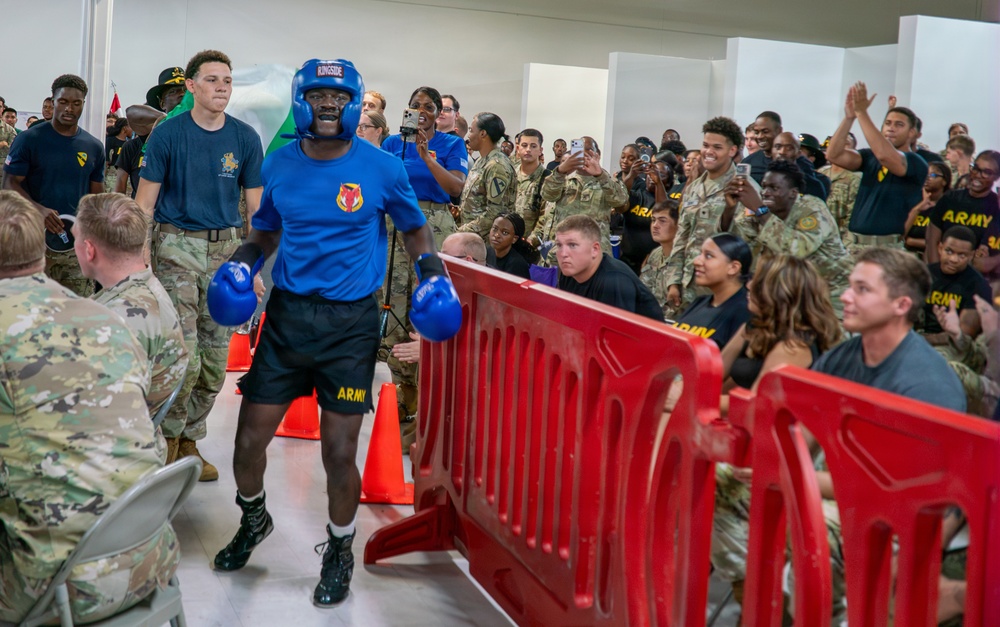 A U.S. Army boxer wearing a blue 'ARMY' uniform, blue boxing gloves, and a blue Ringside headguard walks through a crowd corridor toward a boxing ring or event area. The event appears to be a military boxing match or competition held indoors, with a large crowd of soldiers in camouflage uniforms and civilians cheering enthusiastically. Red crowd-control barriers and orange traffic cones line the walkway.