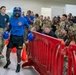 A U.S. Army boxer wearing a blue 'ARMY' uniform, blue boxing gloves, and a blue Ringside headguard walks through a crowd corridor toward a boxing ring or event area. The event appears to be a military boxing match or competition held indoors, with a large crowd of soldiers in camouflage uniforms and civilians cheering enthusiastically. Red crowd-control barriers and orange traffic cones line the walkway.