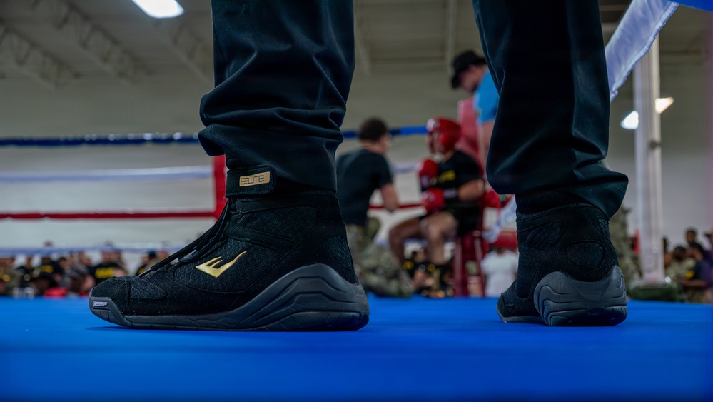 A close-up shot of black and gold Everlast Elite boxing boots standing on a blue boxing ring canvas. The foreground subject is in sharp focus while the background shows a boxing gym environment with fighters and spectators. The image appears to be taken at a boxing event or training session.