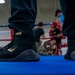 A close-up shot of black and gold Everlast Elite boxing boots standing on a blue boxing ring canvas. The foreground subject is in sharp focus while the background shows a boxing gym environment with fighters and spectators. The image appears to be taken at a boxing event or training session.