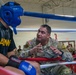 A U.S. Army soldier in camouflage uniform is coaching or giving instructions to a boxer wearing a blue protective headgear and Ringside boxing gloves inside a boxing ring. The boxer is wearing an 'ARMY' branded black shirt, suggesting this is a military boxing event or competition. A crowd of soldiers and personnel can be seen watching in the background.