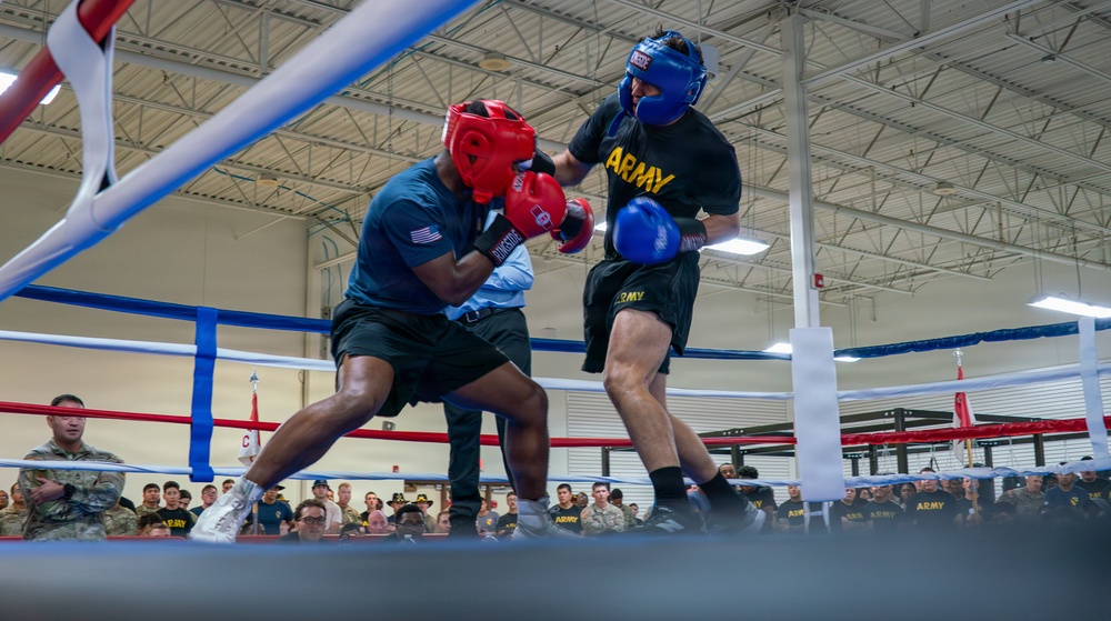Two U.S. Army soldiers are competing in a boxing match inside an indoor boxing ring. One boxer wears red headgear and red gloves, while the other wears blue headgear and blue gloves, both dressed in Army-branded athletic gear. A large crowd of military personnel and spectators watches from ringside in what appears to be a military gymnasium or event facility.