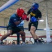 Two U.S. Army soldiers are competing in a boxing match inside an indoor boxing ring. One boxer wears red headgear and red gloves, while the other wears blue headgear and blue gloves, both dressed in Army-branded athletic gear. A large crowd of military personnel and spectators watches from ringside in what appears to be a military gymnasium or event facility.