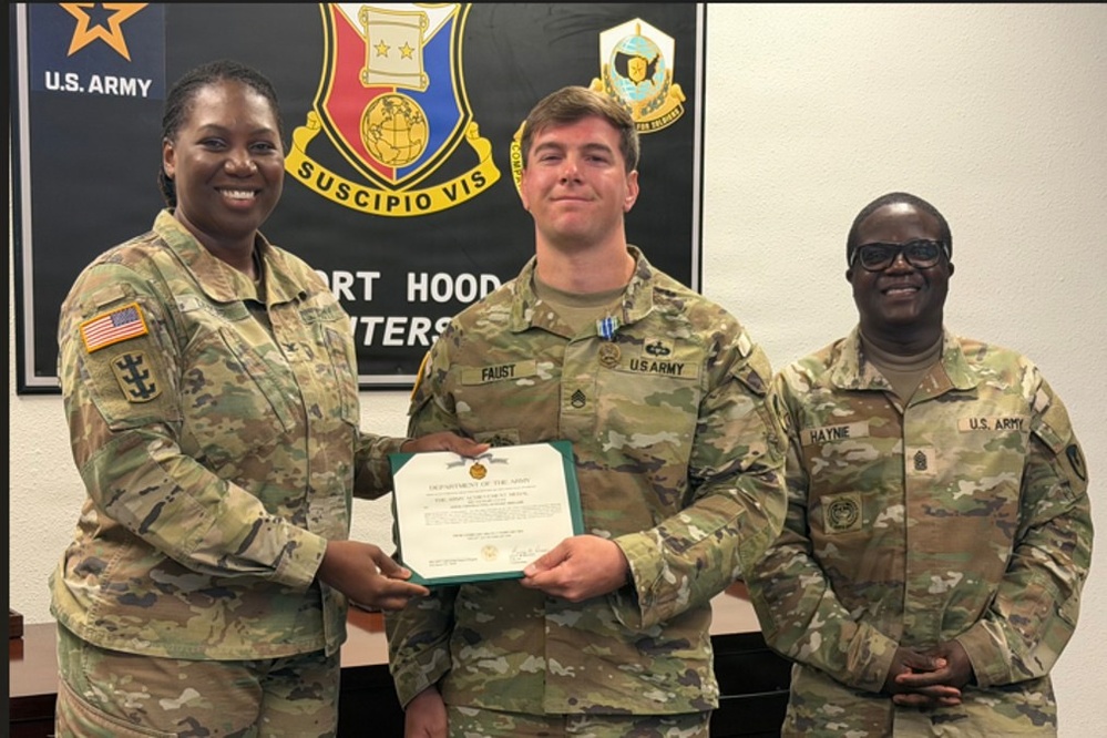 Three U.S. Army soldiers are posing for a photo at Fort Hood headquarters. The center soldier, identified by nametape as 'FAUST,' is receiving a Department of the Army certificate, likely an award or commendation. The soldier on the right is identified by nametape as 'HAYNIE.'