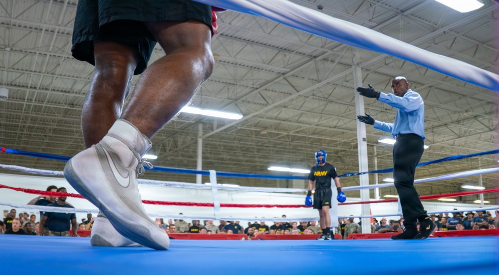 A boxing match is taking place inside a military facility, likely an Army gymnasium, with a large crowd of soldiers watching from ringside. The image is taken from a low angle near the canvas, showing a boxer in white Nike high-tops in the foreground, a referee in a blue shirt separating or signaling, and another boxer wearing an 'ARMY' shirt and blue headgear in the background. The setting and attire suggest this is a military boxing event or competition.