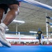 A boxing match is taking place inside a military facility, likely an Army gymnasium, with a large crowd of soldiers watching from ringside. The image is taken from a low angle near the canvas, showing a boxer in white Nike high-tops in the foreground, a referee in a blue shirt separating or signaling, and another boxer wearing an 'ARMY' shirt and blue headgear in the background. The setting and attire suggest this is a military boxing event or competition.