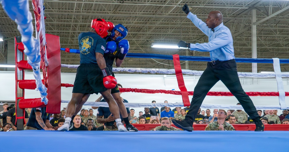 A boxing match is taking place inside a military facility, with two fighters in protective headgear and gloves engaged in close combat. A referee in a light blue shirt and black pants is actively intervening or signaling during the bout. A large crowd of what appears to be military personnel in camouflage uniforms watches from ringside.