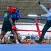A boxing match is taking place inside a military facility, with two fighters in protective headgear and gloves engaged in close combat. A referee in a light blue shirt and black pants is actively intervening or signaling during the bout. A large crowd of what appears to be military personnel in camouflage uniforms watches from ringside.