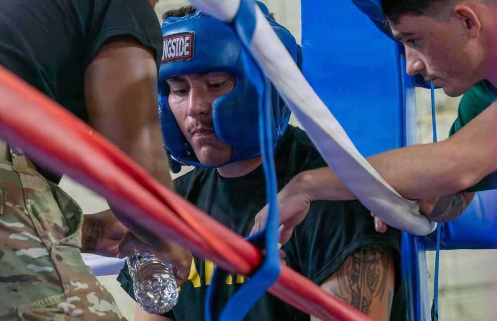 A boxer wearing a blue 'Ringside' brand headgear is in their corner between rounds, receiving attention from corner team members. The fighter appears fatigued or focused, with visible sweat, while cornermen tend to them near the ring ropes. One person in military camouflage pants is visible on the left, suggesting a military boxing event.