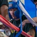 A boxer wearing a blue 'Ringside' brand headgear is in their corner between rounds, receiving attention from corner team members. The fighter appears fatigued or focused, with visible sweat, while cornermen tend to them near the ring ropes. One person in military camouflage pants is visible on the left, suggesting a military boxing event.