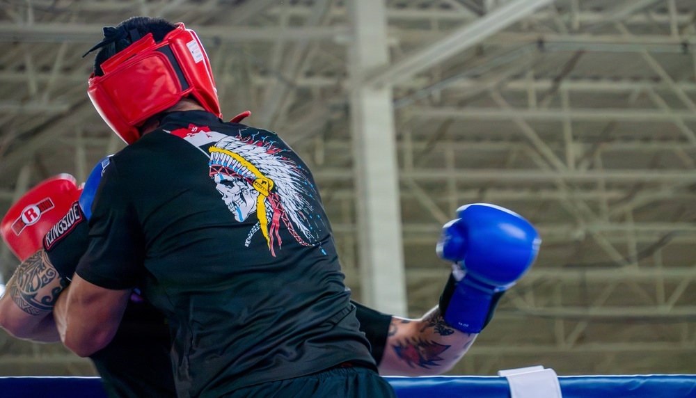 Two boxers are sparring inside a boxing ring in what appears to be a large indoor facility or gymnasium. The fighter facing away from the camera wears a red headguard and red Ringside boxing gloves, along with a black t-shirt featuring a skull with a Native American headdress graphic. The opponent is visible only partially, wearing blue boxing gloves and showing tattooed arms.