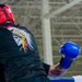 Two boxers are sparring inside a boxing ring in what appears to be a large indoor facility or gymnasium. The fighter facing away from the camera wears a red headguard and red Ringside boxing gloves, along with a black t-shirt featuring a skull with a Native American headdress graphic. The opponent is visible only partially, wearing blue boxing gloves and showing tattooed arms.