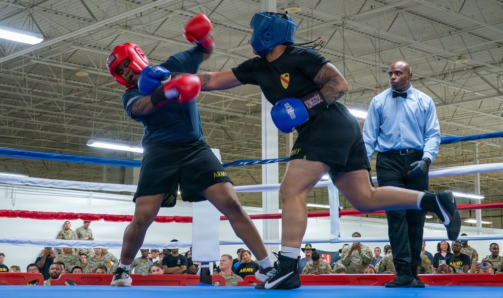 Two U.S. Army soldiers are engaged in a boxing match inside an indoor facility, wearing protective headgear and gloves. A referee in formal attire monitors the bout closely while a large crowd of uniformed military personnel watches from ringside. The match appears to be an organized military boxing event, with both fighters wearing Army-branded shorts and gear.