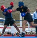 Two U.S. Army soldiers are engaged in a boxing match inside an indoor facility, wearing protective headgear and gloves. A referee in formal attire monitors the bout closely while a large crowd of uniformed military personnel watches from ringside. The match appears to be an organized military boxing event, with both fighters wearing Army-branded shorts and gear.