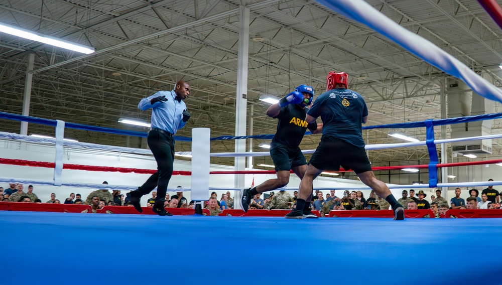 A boxing match is taking place inside a large indoor facility, with two fighters wearing Army-branded gear engaged in an exchange of punches. A referee in a light blue shirt and black pants is actively monitoring the bout at close range. A large crowd of spectators, many appearing to be in military uniforms, watches from the background.