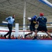 A boxing match is taking place inside a large indoor facility, with two fighters wearing Army-branded gear engaged in an exchange of punches. A referee in a light blue shirt and black pants is actively monitoring the bout at close range. A large crowd of spectators, many appearing to be in military uniforms, watches from the background.