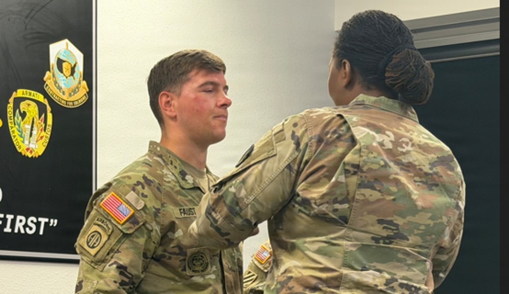 Two U.S. Army soldiers in OCP (Operational Camouflage Pattern) uniforms are engaged in what appears to be a pinning ceremony. A female soldier, seen from behind, is pinning something onto the uniform of a male soldier identified by his name tape as 'FAUST'. The setting appears to be an indoor military facility with unit insignia displayed on the wall.