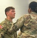 Two U.S. Army soldiers in OCP (Operational Camouflage Pattern) uniforms are engaged in what appears to be a pinning ceremony. A female soldier, seen from behind, is pinning something onto the uniform of a male soldier identified by his name tape as 'FAUST'. The setting appears to be an indoor military facility with unit insignia displayed on the wall.