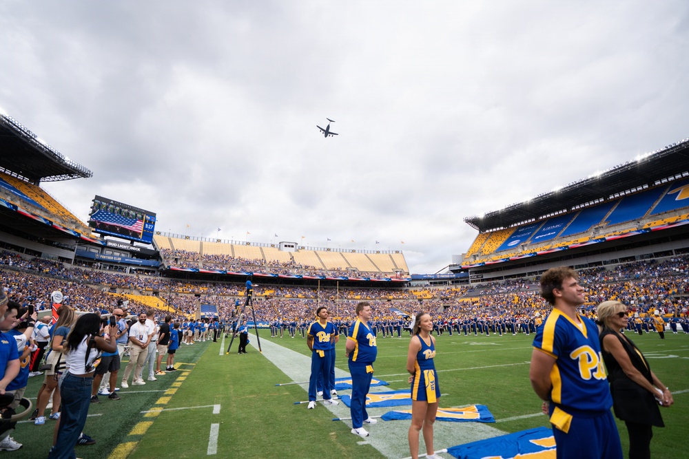 A University of Pittsburgh (Pitt) Panthers football game pregame ceremony is taking place at Acrisure Stadium. Cheerleaders and band members in blue and gold uniforms stand on the field while a military aircraft performs a flyover above the stadium. The stands are filled with fans, and an American flag is visible on the large video scoreboard.