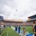 A University of Pittsburgh (Pitt) Panthers football game pregame ceremony is taking place at Acrisure Stadium. Cheerleaders and band members in blue and gold uniforms stand on the field while a military aircraft performs a flyover above the stadium. The stands are filled with fans, and an American flag is visible on the large video scoreboard.