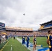 A University of Pittsburgh (Pitt) Panthers football game pregame ceremony is taking place at Acrisure Stadium. Cheerleaders and band members in blue and gold uniforms stand on the field while a military aircraft performs a flyover above the stadium. The stands are filled with fans, and an American flag is visible on the large video scoreboard.
