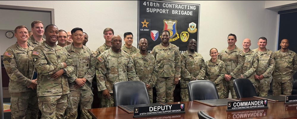 A group of approximately 20 U.S. Army soldiers in OCP (Operational Camouflage Pattern) uniforms pose for a formal group photo inside a conference room of the 418th Contracting Support Brigade. The setting includes a large unit insignia display board on the wall and a conference table with nameplates for 'Deputy' and 'Commander' of the 418th Contracting Support Brigade. The group is diverse in rank, gender, and ethnicity.