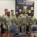 A group of approximately 20 U.S. Army soldiers in OCP (Operational Camouflage Pattern) uniforms pose for a formal group photo inside a conference room of the 418th Contracting Support Brigade. The setting includes a large unit insignia display board on the wall and a conference table with nameplates for 'Deputy' and 'Commander' of the 418th Contracting Support Brigade. The group is diverse in rank, gender, and ethnicity.