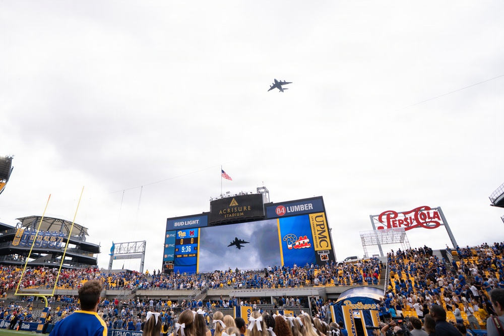 A military flyover is taking place above Acrisure Stadium during what appears to be a college football game, likely a University of Pittsburgh (Pitt Panthers) home game. The large aircraft is visible in the sky above the stadium while the jumbotron displays a live feed of the flyover. Cheerleaders, fans in blue and gold colors, and stadium signage are prominently visible.