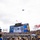 A military flyover is taking place above Acrisure Stadium during what appears to be a college football game, likely a University of Pittsburgh (Pitt Panthers) home game. The large aircraft is visible in the sky above the stadium while the jumbotron displays a live feed of the flyover. Cheerleaders, fans in blue and gold colors, and stadium signage are prominently visible.