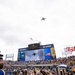A military flyover is taking place above Acrisure Stadium during what appears to be a college football game, likely a University of Pittsburgh (Pitt Panthers) home game. The large aircraft is visible in the sky above the stadium while the jumbotron displays a live feed of the flyover. Cheerleaders, fans in blue and gold colors, and stadium signage are prominently visible.