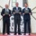 Three men in business suits are posing for a formal photograph while holding black certificate folders branded with 'Leadership Pittsburgh Inc.' They appear to be at a graduation or recognition ceremony, standing on a red platform in front of a geometric white wall structure. The setting appears to be an indoor venue with decorative ivy planters on either side.