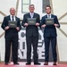 Three men in business suits are posing for a formal photograph while holding black certificate folders branded with 'Leadership Pittsburgh Inc.' They appear to be at a graduation or recognition ceremony, standing on a red platform in front of a geometric white wall structure. The setting appears to be an indoor venue with decorative ivy planters on either side.