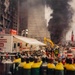 This image depicts an active disaster response operation at Ground Zero following the September 11, 2001 attacks in New York City. Emergency personnel, heavy equipment, and command vehicles are visible amid the rubble, with large plumes of dark smoke rising in the background. Multiple agency resources including US Army Corps of Engineers Emergency Operations units are deployed at the scene.