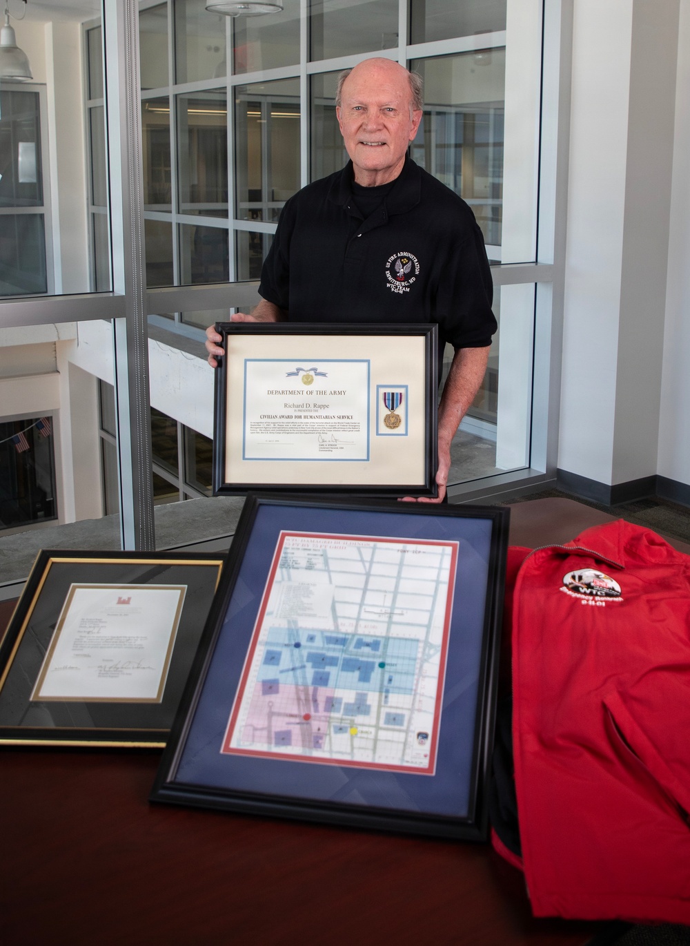 An older man wearing a black polo shirt stands behind a table displaying several framed awards and memorabilia. He is holding up a framed Department of the Army 'Civilian Award for Humanitarian Service' certificate issued to Richard D. Rappe, which includes a medal. Additional framed documents, a colored map, and a red jacket are displayed on the table in front of him.