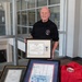 An older man wearing a black polo shirt stands behind a table displaying several framed awards and memorabilia. He is holding up a framed Department of the Army 'Civilian Award for Humanitarian Service' certificate issued to Richard D. Rappe, which includes a medal. Additional framed documents, a colored map, and a red jacket are displayed on the table in front of him.