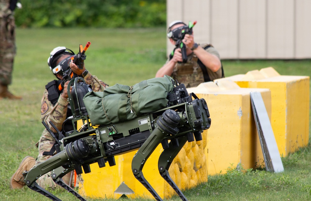 Two military personnel in camouflage uniforms are conducting a training exercise with a quadruped robot dog, likely a Boston Dynamics Spot or similar platform, which is carrying military gear. The soldiers are using training weapons with orange/red barrel blockers and wearing protective eyewear, indicating a simulated combat drill. Yellow concrete barriers serve as cover positions during the exercise.