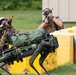 Two military personnel in camouflage uniforms are conducting a training exercise with a quadruped robot dog, likely a Boston Dynamics Spot or similar platform, which is carrying military gear. The soldiers are using training weapons with orange/red barrel blockers and wearing protective eyewear, indicating a simulated combat drill. Yellow concrete barriers serve as cover positions during the exercise.