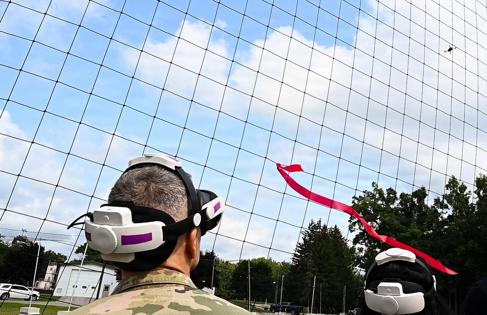 Two individuals wearing VR/AR headsets are observing what appears to be a small drone or UAV flying within a large netted enclosure outdoors. A red ribbon or streamer is visible in the air, possibly attached to the drone or used as a visual marker. The setting appears to be a military or research facility based on the camouflage uniform visible on one person.