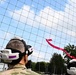 Two individuals wearing VR/AR headsets are observing what appears to be a small drone or UAV flying within a large netted enclosure outdoors. A red ribbon or streamer is visible in the air, possibly attached to the drone or used as a visual marker. The setting appears to be a military or research facility based on the camouflage uniform visible on one person.