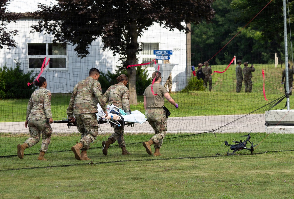 Military personnel in camouflage uniforms are carrying a patient on a stretcher during what appears to be a medical training exercise. Red ribbon markers are visible throughout the scene, and a drone is on the ground near a net barrier. A building marked 'HOSPITAL' is visible in the background with additional uniformed personnel.