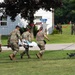 Military personnel in camouflage uniforms are carrying a patient on a stretcher during what appears to be a medical training exercise. Red ribbon markers are visible throughout the scene, and a drone is on the ground near a net barrier. A building marked 'HOSPITAL' is visible in the background with additional uniformed personnel.