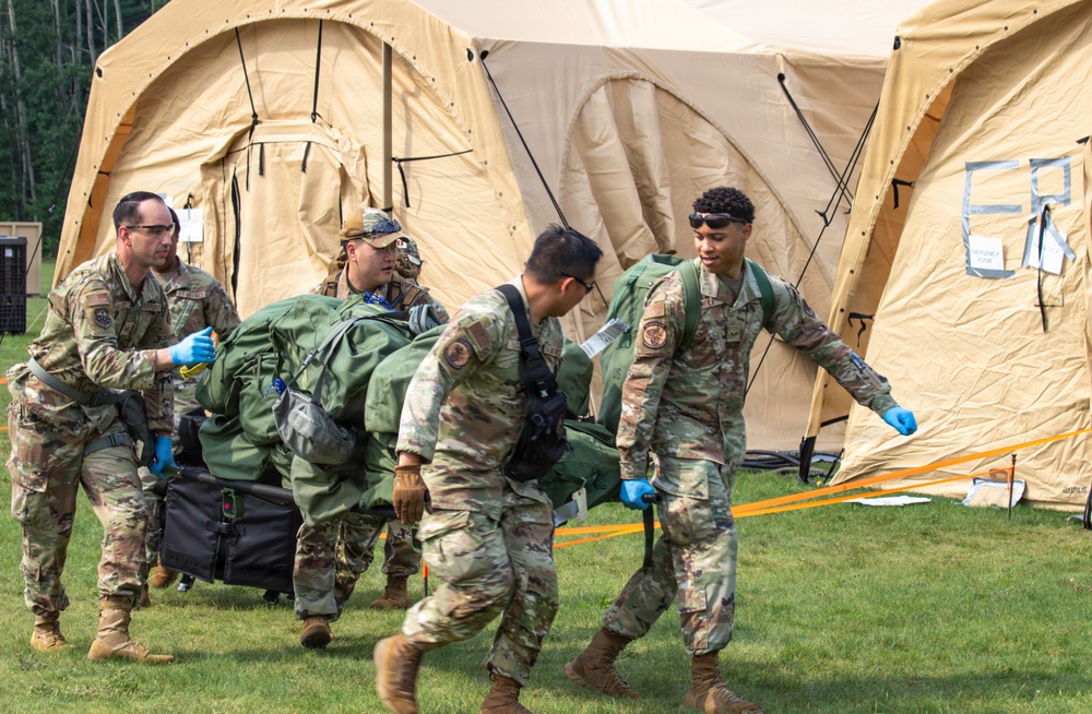 Military personnel in camouflage uniforms are carrying a patient on a litter/stretcher toward large tan military field medical tents. The scene appears to be a field medical exercise or training scenario, with personnel wearing blue medical gloves indicating a medical response context. Multiple service members are coordinating the casualty transport in an outdoor grassy area.